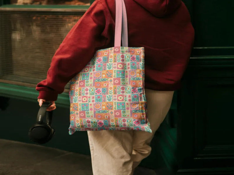 A person wearing a burgundy hoodie and beige pants carries a colorful patterned canvas tote bag with pink shoulder straps, while holding a black headphones in one hand, set against a dark green wooden door background.